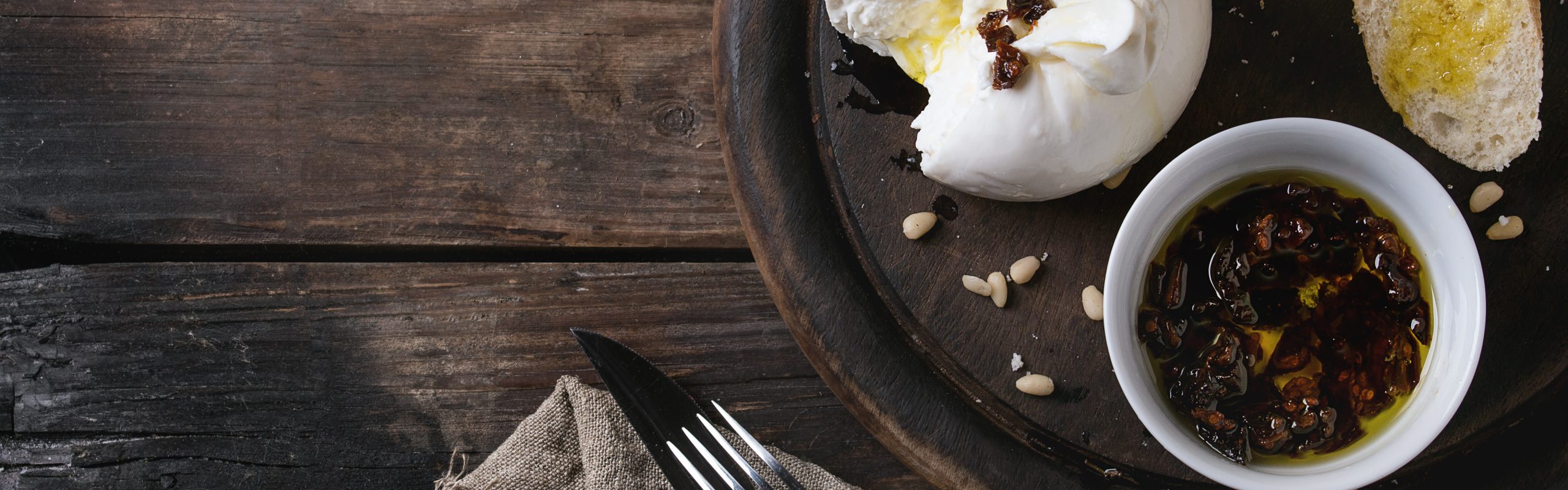 Sliced Italian cheese burrata served on round chopping board with pine nuts, sliced bread, basil and dried tomatoes in olive oil with fork and knife over old wood background. Overhead view